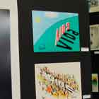 Nesta foto em cores observa-se painel com três cartuns expostos. Um deles mostra pessoas segurando nos calcanhares da outra, de maneira a formarem uma corrente para alcançar uma pessoa sentada no fundo do poço. Nota-se que em cada indivíduo há o símbolo de negativo ou positivo. Em outro cartum, nota-se a palavra Aids e sua sombra, que forma a palavra vida.