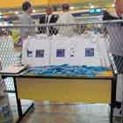 Nesta foto em cores e em close, veem-se mesa repleta de embalagens de camisinhas e sacolas. Nas laterais, porta fôlderes com material impresso e colorido. Ao fundo, observam-se algumas pessoas.