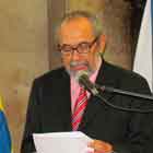 Esta foto em cores apresenta um senhor falando ao microfone. Percebe-se que ele está em um púlpito e o microfone em pedestal. Atrás dele, notam-se duas bandeiras, sendo uma do Brasil.