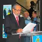 Nesta foto em cores, um senhor está em pé atrás de um púlpito, falando ao microfone. Ao fundo, notam-se painéis preenchidos com cartuns, além de algumas pessoas em pé entre as estruturas. 