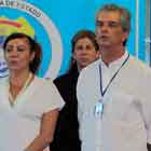 Nesta fotografia em cores, observam-se duas mulheres e um homem. Percebe-se a expressão séria nos rostos deles e nota-se que o homem tem um crachá no pescoço. Ao fundo, parte do brasão de uma secretaria de estado e a bandeira do Brasil.