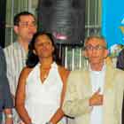 Esta foto em cores revela grupo de pessoas em pé, lado a lado e de frente para a câmera. Percebe-se que não olham para o fotógrafo e um senhor coloca a mão direita no peito, sob o paletó.