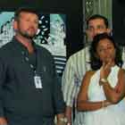 Quatro pessoas aparecem nesta foto. Elas estão em pé, lado a lado, cada uma olhando para uma direção. Nota-se que atrás deles há uma estrutura vertical preta onde foram afixados alguns cartuns.