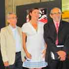 Na foto em cores, dois homens e uma mulher posam para a foto. Estão em pé ao lado de banner do I Festival Internacional de Humor em DST e Aids. Ainda no banner, observa-se o desenho de dois corações sorridentes dentro de uma camisinha. Ao fundo, notam-se estruturas verticais pretas com cartuns afixados. 