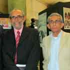 Três senhores posam para a câmera nesta foto em cores. Apenas um deles, à direita, não sorri. Percebem-se, ao fundo, estruturas verticais pretas nas quais foram afixados cartuns. 