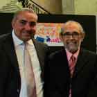 Um mulher e três senhores posam para a câmera nesta foto em cores. Apenas um deles, à direita, não sorri. Percebem-se, ao fundo, estruturas verticais pretas nas quais foram afixados cartuns.