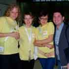 Nesta foto, três mulheres de camiseta amarela estão em pé, lado a lado e acompanhadas por um rapaz de terno. Percebe-se que todos posam para a câmera e sorriem.