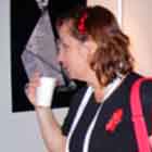 Uma visitante da mostra aparece nesta foto. Nota-se que ela olha para o painel onde estão os cartuns enquanto toma algo em um copo descartável. Percebe-se que ela usa broche com o símbolo da luta contra a Aids, um laço vermelho.