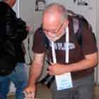 Um senhor inclina o tronco para frente a fim de escrevem em caderno aberto sobre mesa repleta de fôlderes coloridos. A foto mostra também outras pessoas observando os cartuns expostos ao fundo.