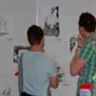 Nesta foto em cores, dois rapazes param diante de alguns cartuns expostos e observam. À direita, vê-se outro homem em pé, de braços cruzados sobre o peito.