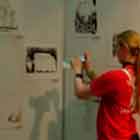 Esta foto mostra uma jovem em pé em frente a alguns cartuns. Percebe-se que ela fotografa o painel.