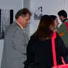 Nesta foto em cores, um homem e uma mulher param em frente à mesa repleta de fôlderes. Notam-se outras pessoas ao fundo, observando catálogo impresso ou cartuns expostos nos painéis.