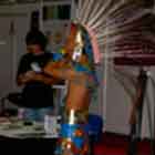 Esta foto mostra um homem trajado com adereços indígena e usando grande cocar na cabeça. Nota-se que ele conversa com um visitante da mostra. Ao fundo, painéis brancos abrigam itens expostos.