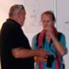 Nesta foto, à esquerda, uma mulher observa alguns cartuns afixados num painel branco. À direita, nota-se que um homem conversa com uma mulher que leva uma câmera presa ao pescoço.