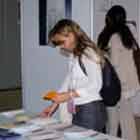  Nesta foto, vê-se uma moça escolhendo fôlder entre os materiais impressos e disponibilizados sobre a mesa. Ao fundo, notam-se algumas pessoas observando os painéis da mostra.