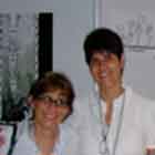 Nesta foto, duas mulheres estão lado a lado, abraçadas, posando e sorrindo para a câmera. Nota-se, ao fundo, painel com cartuns em preto e branco.