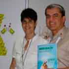 Nesta foto em cores, mulher e homem, lado a lado posam e sorriem para a câmera. Nota-se que o homem mostra publicação cujo título diz “More Health”. Ao fundo, notam-se dois cartuns expostos no painel.