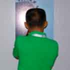 Dois homens observam os cartuns expostos nos painéis nesta foto em cores. Percebe-se que um deles fotografa alguma obra.