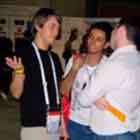 Nesta foto, três rapazes conversam ao lado de uma mesa repleta de materiais impressos. Percebe-se que um dos jovens gesticula enquanto fala. Ao fundo, notam-se painéis alguns painéis e pessoas circulando pelo local.