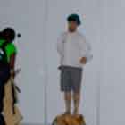 Esta foto mostra pessoas passando, caminhando para a esquerda ou para a direita. Nota-se que no centro há um rapaz em pé e parado sobre um degrau. Percebe-se ainda que algumas pessoas param para vê-lo.