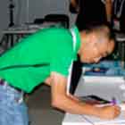 Um homem inclina o tronco para frente a fim de escrever em caderno aberto sobre mesa repleta de fôlderes. A foto em cores mostra também um rapaz em pé, atrás da mesa, observando o visitante.