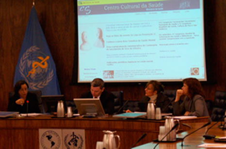 Esta fotografia em cores mostra quatro pessoas sentadas atrás de bancada que possui computador e microfones de mesa. Atrás deles, um telão mostra imagem sobre o Centro Cultural da Saúde, hoje Centro Cultural do Ministério da Saúde. Percebe-se que a mulher à esquerda é quem fala ao microfone. Ao fundo, nota-se a bandeira da Organização Mundial de Saúde (OMS), que é uma bandeira azul com o símbolo da medicina em amarelo sobreposto a um desenho de mapa mundi rodeado de ramos de oliveira.