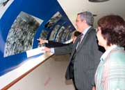 A fotografia mostra, de perfil, mulher e homem de terno que aponta para foto em preto e branco exposta junto a uma parede curva, à esquerda. Nota-se que outra pessoa, ao lado do homem, também aponta para a mesma foto. Percebe-se ainda que, ao longo do corredor, estão expostas muitas outras fotografias.