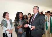 Esta foto em cores revela pessoas em volta de um homem que gesticula enquanto fala. Nota-se que todos olham para ele e que estão em local fechado e claro.