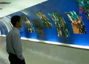 Esta foto mostra um homem em pé, com as mãos nos bolsos, olhando para as fotos a sua frente. Percebe-se que foram impressas em fundo azul afixado em toda a parede de um corredor.