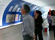 Esta foto mostra um homem em pé, com as mãos para trás, olhando para as fotos a sua frente. Além dele, outras pessoas também observam a mostra. Percebe-se que as imagens expostas foram impressas em fundo azul afixado em toda a parede de um corredor.