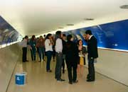 Esta foto colorida mostra um corredor que, na verdade é um túnel, ocupado nas laterais por uma exposição. Notam-se fotos e outras imagens impressas em fundo azul ao longo de todo o caminho. Percebe-se também que as pessoas param para observar a mostra.