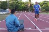 Not�cia: �Tecnologia permite que atletas cegos corram sozinhos�. Descri��o da imagem: a fotografia mostra uma pista de corrida em um campo de treinamento, com a devida pavimenta��o e as marca��es das raias. Em primeiro plano, h� um homem sentado, de costas para a c�mera, usando um computador do tipo notebook. O homem est� voltado para dois corredores que est�o juntos em uma s� raia. Destes dois homens que est�o na pista, vestidos com roupa e t�nis pr�prios para corrida, nota-se que o de tr�s apoia o outro. O corredor da frente usa nos bra�os uma esp�cie de bracelete preto. Fim da descri��o.