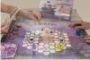 Not�cia: �UnB lan�a jogo que discute viol�ncia contra a mulher�. Descri��o da imagem: fotografia onde se observa um tabuleiro de jogo em tons de roxo e violeta, com uma mandala no centro, composta por v�rios c�rculos brancos interligados, sobre o qual h� pe�es, fichas, dados e cart�es coloridos. De cada lado do tabuleiro, apoiado sobre uma mesa branca, v�-se um par de m�os; o da esquerda, com uma das m�os, segura um cart�o, e com a outra manipula um dos pe�es; o da direita apenas apoia as m�os sobre o tabuleiro. Ao fundo, h� a caixa do jogo, com o desenho de uma silhueta feminina e a palavra �VIOLETAS� escrita em roxo, al�m de outros textos ileg�veis. Fim da descri��o.