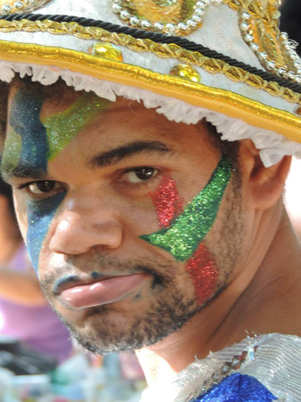 Imagem. A fotografia em formato retrato, isto �, exposta na orienta��o vertical, mostra um foli�o que, virando o rosto � esquerda, encara a lente da c�mera. O rosto do fotografado ocupa a imagem quase inteiramente. O foli�o � um jovem branco, de aproximadamente 30 anos de idade. Ele tem barbas por fazer. Todo o rosto est� pintado com faixas que, nas cores verde, vermelho e azul, desenham tra�os abstratos em sua pele. Seus olhos s�o pretos, bastante escuros. Sua express�o � s�ria, com o semblante muito fechado. Ele usa um chap�u que foi empregado, provavelmente, em um desfile de escola de samba. O adere�o � branco com detalhes dourados, pretos e um cord�o com bolinhas prateadas. O fundo da imagem � composto por formas e cores indistintas, de partes das fantasias de outros foli�es. Foto: �rike de Freitas. Fim da descri��o.