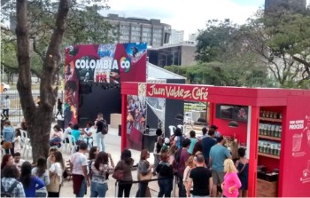 Segunda imagem: Fotografia onde se v,  esquerda, um stand vermelho, na parte de cima do qual se l JUAN VALDEZ CAFɔ, em branco. Em frente ao stand, h uma pequena aglomerao de pessoas, seguida por uma fila. Atrs da fila  possvel ver o tronco de uma rvore grande, que se bifurca em dois, e, atrs da rvore, vrias cadeiras de plstico brancas, onde algumas pessoas esto sentadas. Em frente a essas cadeiras, ao fundo do stand, v-se um palco, onde se l COLOMBIA CO. Ao fundo, rvores, prdios e nuvens. Fim da descrio.