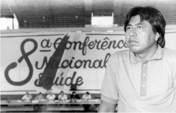 Galeria de imagens. Primeira imagem: Nesta foto em preto e branco, v-se um homem indgena  esquerda. Ao fundo, nota-se um grande painel escrito 8 Conferncia Nacional de Sade. Logo abaixo, percebe-se uma grande bancada com pessoas sentadas, como se fossem palestrar. Acervo: Departamento de Arquivo e Documentao/Casa de Oswaldo Cruz/Fiocruz. Fotgrafo: lvaro Pedreira. Fim da descrio.