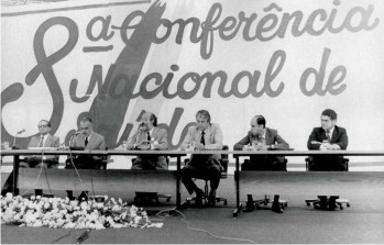 Segunda imagem: Nesta foto em preto e branco, v-se uma grande bancada com pessoas sentadas, como se fossem palestrar. Ao fundo, nota-se um grande painel escrito 8 Conferncia Nacional de Sade. Fim da descrio.