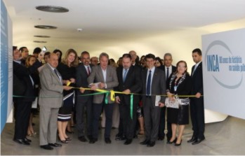 Segunda imagem: Foto do momento de descerramento da faixa inaugural da uma exposio, que acontece no tnel que liga a sede do Ministrio da Sade ao prdio anexo, em Braslia. Uma fita de tecido verde e amarelo est disposta entre dois grandes murais que compem a exposio. Um homem em trajes formais, vestindo um palet cinza segura uma das pontas da fita e  ajudado por mais dois homens e uma mulher ao lado esquerdo. Do seu lado direito, um outro homem segura outra parte da fita. Nas laterais e ao fundo h vrios homens e mulheres que acompanham o ato inaugural. Fim da descrio.