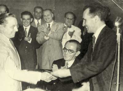 Em preto e branco, a fotografia mostra o cumprimento entre dois homens. Em primeiro plano, cada um em uma lateral da imagem, os homens cumprimentam-se com um aperto de mãos e sorriem. Entre eles, há um homem sentado e, atrás desse, vários outros estão em pé, aplaudindo. 
