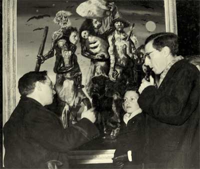 A fotografia, em preto e branco, retrata pessoas conversando em frente a uma das pinturas de Portinari, intitulada “Os retirantes”. Há um homem à esquerda, de perfil, conversando e gesticulando com a mão direita. Ele se dirige a uma mulher e a um homem, também posicionados de perfil.