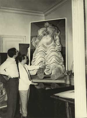 Na fotografia, em preto e branco, dois homens observam um quadro de Portinari. Estão em pé, à esquerda. O homem mais perto da tela estica o braço esquerdo até quase tocá-la, enquanto o outro, ao lado dele, apoia a mão direita no quadril e olha para cima. Ao lado direito, percebe-se um recipiente cilíndrico com muitos pincéis dentro, além da quina de uma mesa. Ao fundo, partes de outras obras aparecem afixadas na parede. 