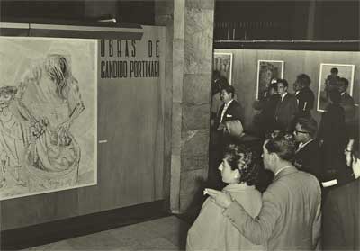 Foto, em preto e branco, mostra visitantes em uma exposição. Na lateral esquerda há uma parede com um painel que reproduz uma pintura do artista e, ao lado, as palavras “Obras de Cândido Portinari”. Na lateral direita, e mais ao fundo, há pessoas circulando entre painéis afixados nas paredes. 