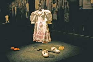 Esta fotografia em cores mostra, em close, vestido de criança pendurado em cabide. A roupa, em cor clara e babados, parece flutuar sobre um piso escuro de carpete, onde se encontram um par de sapatos, também claros, e uma boneca. Ao fundo, nota-se um grande painel com foto em preto e branco que mostra várias pessoas lado a lado, de mãos dadas. Esta fotografia em cores mostra, em close, vestido de criança pendurado em cabide. A roupa, em cor clara e babados, parece flutuar sobre um piso escuro de carpete, onde se encontram um par de sapatos, também claros, e uma boneca. Ao fundo, nota-se um grande painel com foto em preto e branco que mostra várias pessoas lado a lado, de mãos dadas.