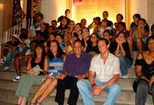 Esta foto em cores mostra grupo de pessoas sentadas em escadaria que dá acesso a um edifício. Posicionado lado a lado, percebe-se que alguns olham para a câmera e sorriem, enquanto outros apresentam uma feição séria.