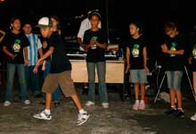 Um grupo de pré-adolescentes está de frente para a câmera. Nota-se que um deles está fora do conjunto e à frente dos demais. O menino em destaque está em movimento, voltado para a esquerda. Usa boné, camiseta preta, bermuda marrom e tênis brancos. Atrás dos integrantes do grupo, observa-se uma mesa e um homem em pé.