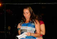 Nesta fotografia em cores, uma mulher destaca-se por estar no centro, em pé e falando ao microfone. Nota-se que ela olha para um pedaço de papel que segura a sua frente. Ao fundo nota-se a presença de outra mulher.