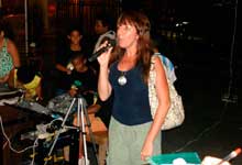 Nesta fotografia em cores, uma mulher destaca-se por estar no centro, em pé e falando ao microfone. Nota-se que ela olha para frente e que leva uma bolsa no ombro esquerdo. Ao fundo nota-se a presença de garotos pré-adolescentes atrás de mesa com equipamento de som.