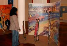 Nesta foto, vê-se mulher em pé, de perfil, observando um dos painéis da mostra. Nota-se que à esquerda há outro painel.