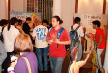 Nesta fotografia colorida, várias pessoas estão juntas em um salão onde há painéis da mostra. Observa-se que os visitantes movimentam-se em diferentes direções ou conversam em pequenos grupos. 