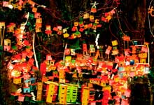 Foto revela maquete na qual se reconhece fração de uma comunidade ou favela que cresceu sobre um morro. As pequenas casas coloridas e prédios foram erguidos sem planejamento, ocupando os contornos do monte. Notam-se muitos fios, também desordenados, representando as ligações caseiras que levam energia elétrica às moradias.